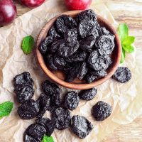 bowl of dried prunes surrounded with more prunes and leaf and plums on the paper at the table