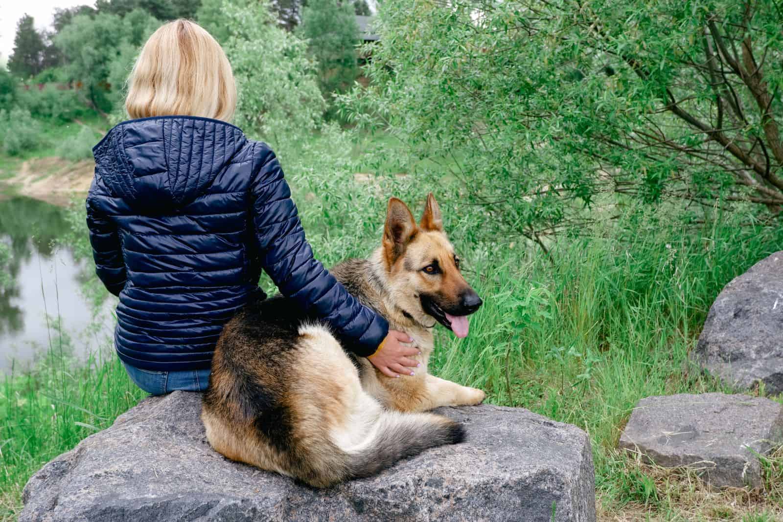 A young woman with a German Shepherd sits on a large granite stone by the lake