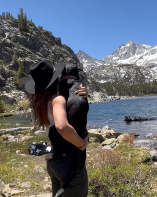 a woman with a hat is standing next to the river and holding a dog in her arms