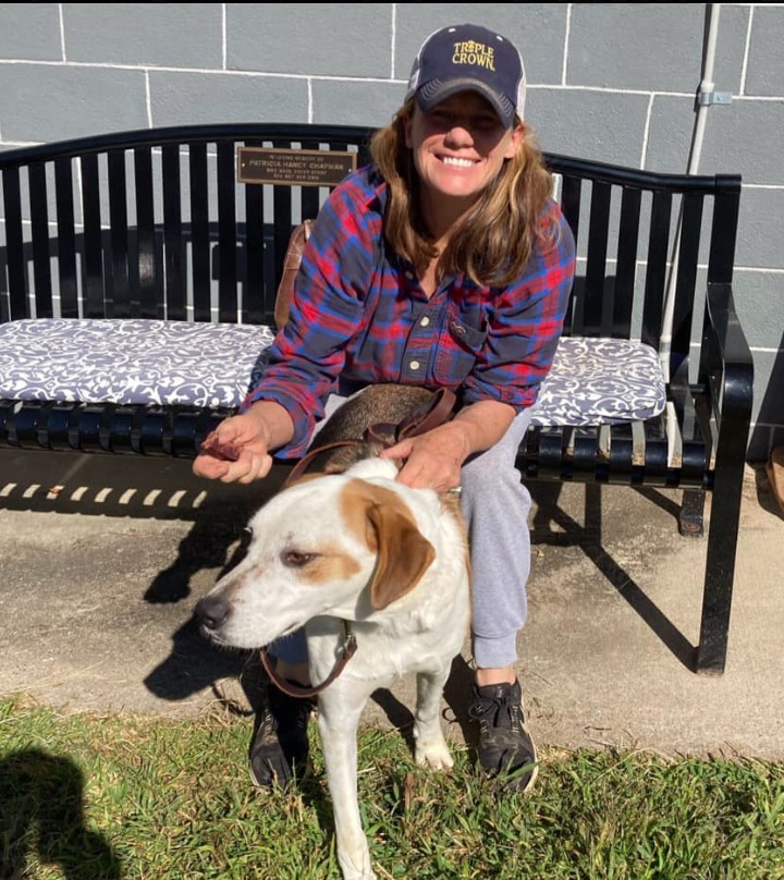 a smiling woman on a park bench holds her dog next to her on a leash